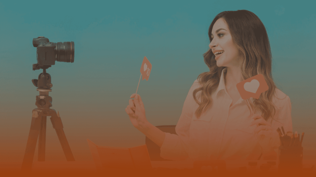 Whissel Strategies A woman sits at a desk in front of a camera on a tripod, holding paper cutouts of social media like and love icons, smiling and looking at the camera. Toronto Digital Marketing Agency