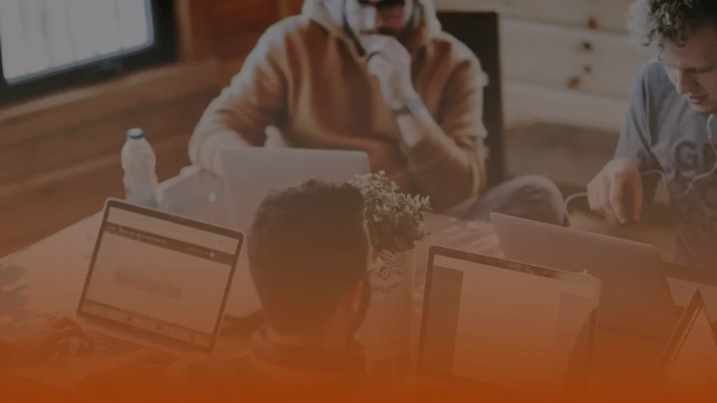 Whissel Strategies Three people work on laptops around a table, each focused on their screens. A potted plant sits in the center of the table. The foreground has an orange overlay. Toronto Digital Marketing Agency