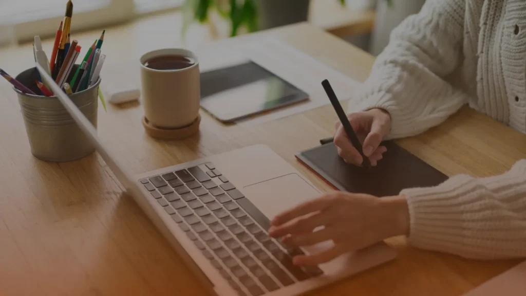Whissel Strategies Person using a laptop and graphic tablet at a wooden desk with a cup of coffee, tablet, and pencils nearby. Toronto Digital Marketing Agency