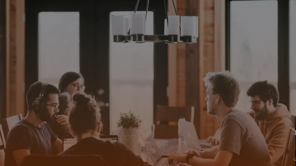 Whissel Strategies Four people sit around a table working on laptops, with a potted plant in the center and a modern chandelier above them in a room with large windows. Toronto Digital Marketing Agency