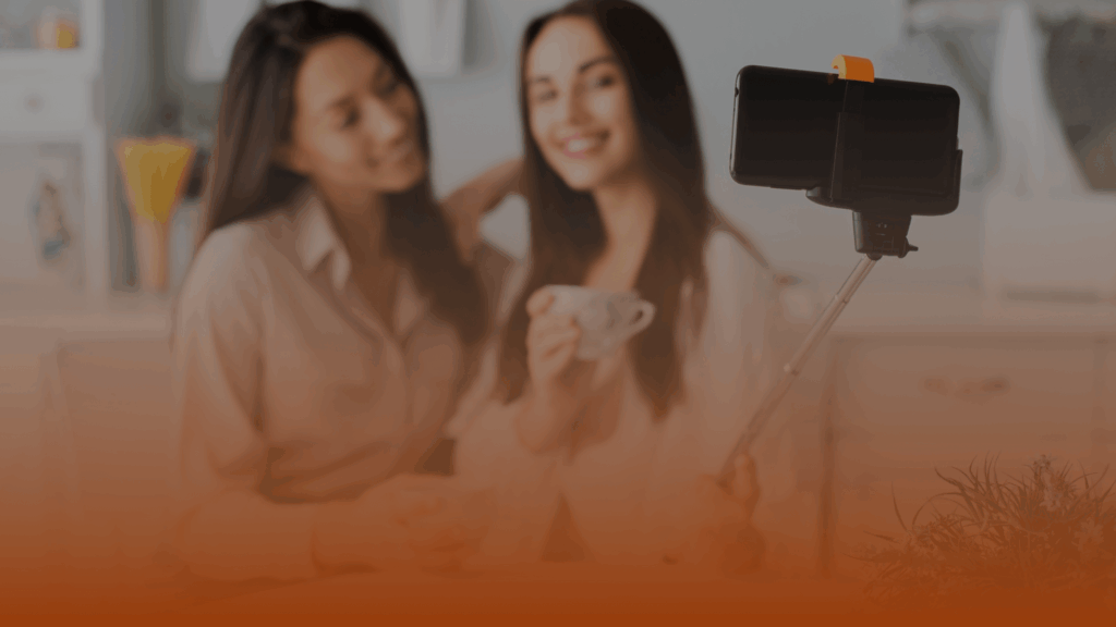 Whissel Strategies Two women sit together indoors, smiling at a smartphone mounted on a selfie stick; one holds a teacup. Toronto Digital Marketing Agency