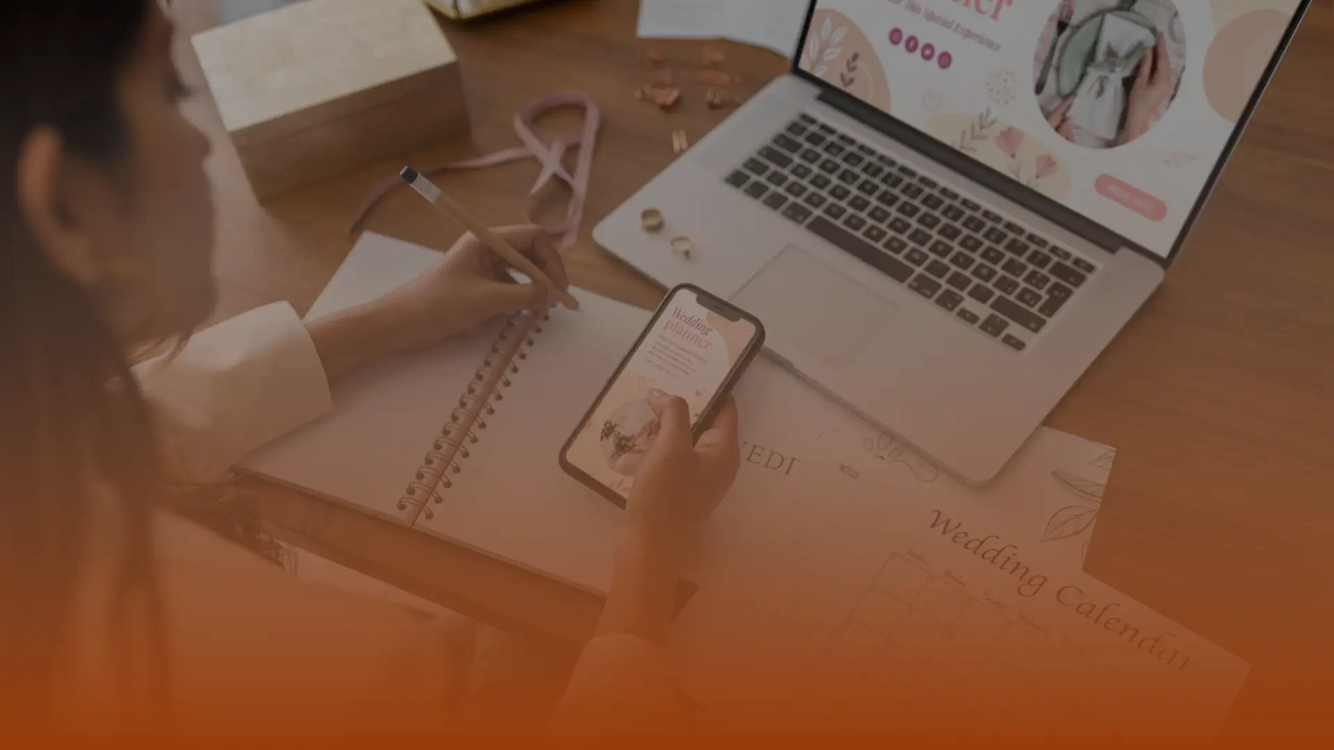 Whissel Strategies Person writing in a notebook while holding a smartphone, with an open laptop and a wedding calendar on the desk. Toronto Digital Marketing Agency