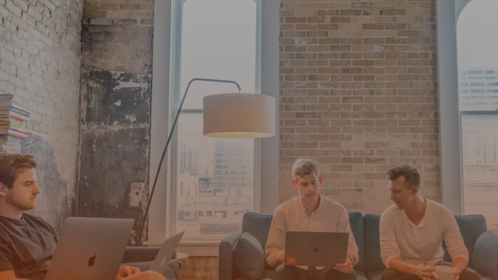 Whissel Strategies Four people sit on couches in a modern office with exposed brick walls, using laptops and talking, with large windows and a floor lamp in the background. Toronto Digital Marketing Agency