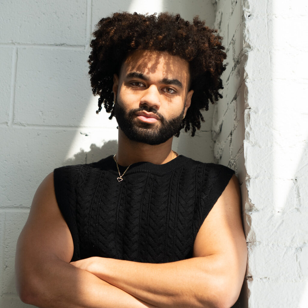 Whissel Strategies A person with curly hair and a beard stands against a white brick wall, arms crossed, wearing a sleeveless black sweater and black pants, lit by natural sunlight. Toronto Digital Marketing Agency