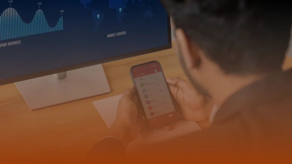 Whissel Strategies A person sits at a desk viewing financial data on a monitor while holding a smartphone displaying a list of items or messages. Toronto Digital Marketing Agency