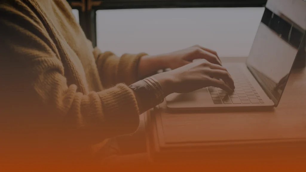 Whissel Strategies Person wearing a sweater types on a laptop at a wooden desk near a window, with an orange gradient overlay at the bottom of the image. Toronto Digital Marketing Agency