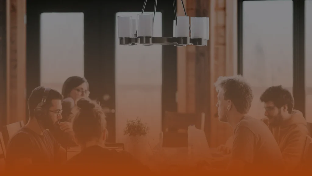 Whissel Strategies Four people sit around a table in a meeting room, engaged in discussion. A modern ceiling light fixture hangs above, and large windows provide natural light. Toronto Digital Marketing Agency