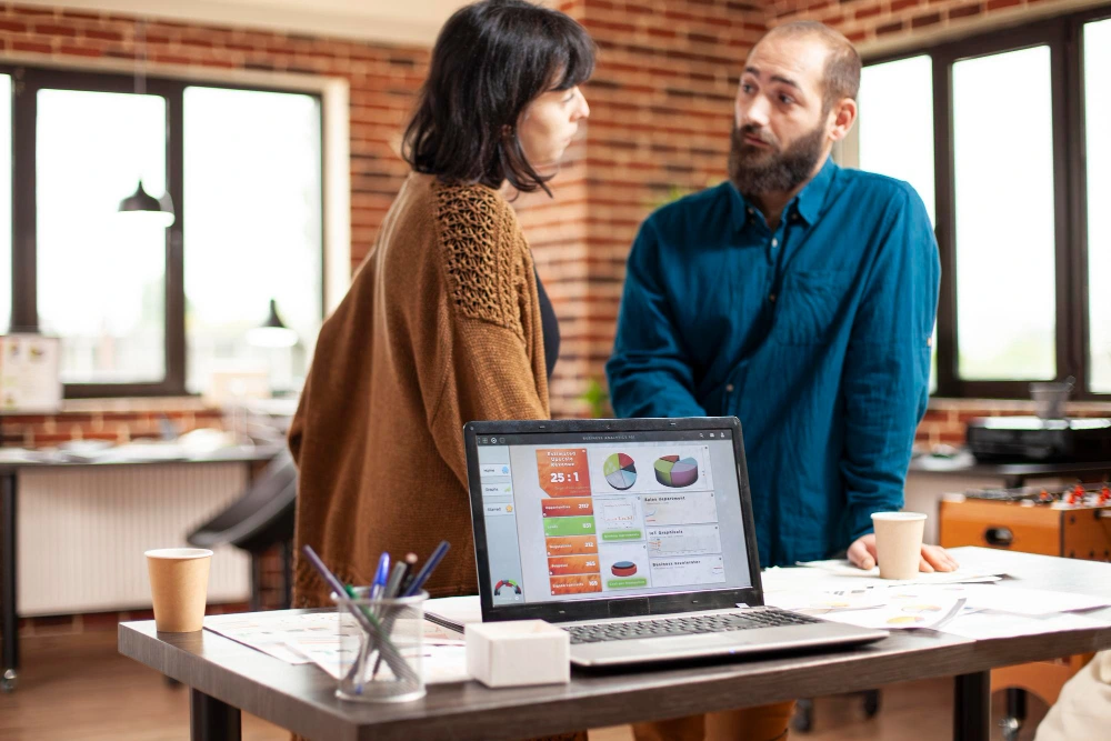Whissel Strategies Two people talk by a desk with a laptop displaying charts and documents. Office supplies and paper cups are on the table in a modern, brick-walled workspace. Toronto Digital Marketing Agency