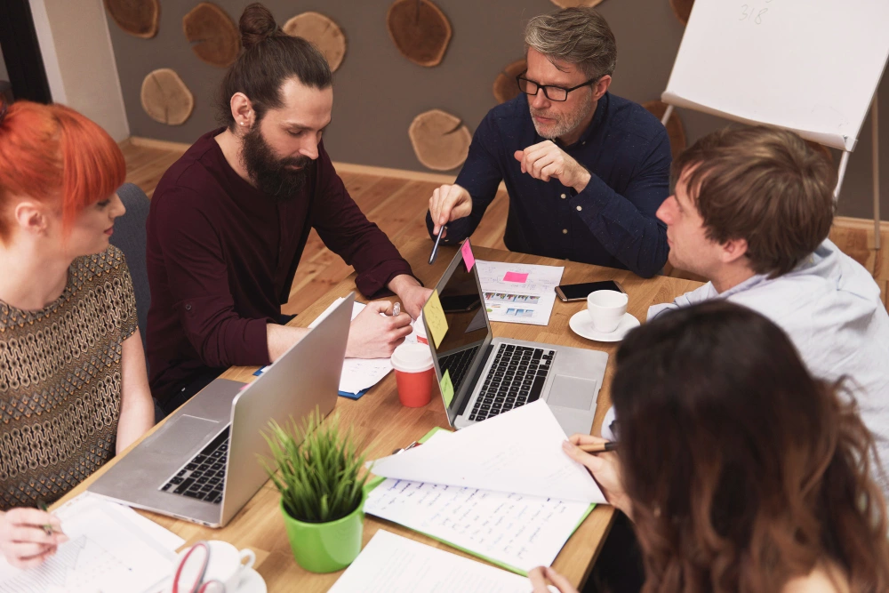 Whissel Strategies Five people sit around a table with laptops, papers, coffee, and a plant, discussing work in a casual office setting with a flipchart in the background. Toronto Digital Marketing Agency