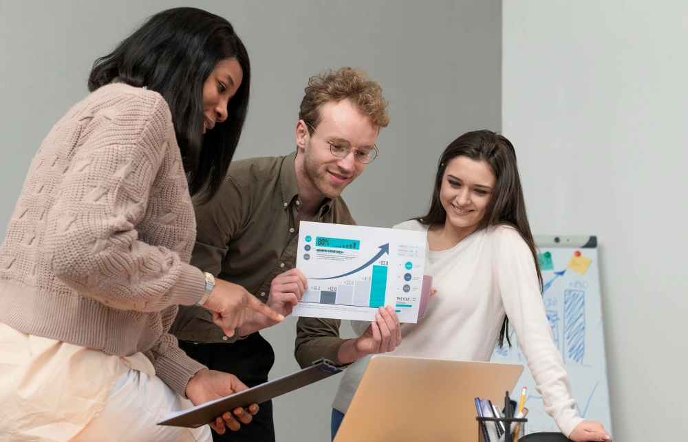 Whissel Strategies Three people collaborate in an office, reviewing a printed chart with graphs and data while smiling and looking at a laptop on a desk. Toronto Digital Marketing Agency