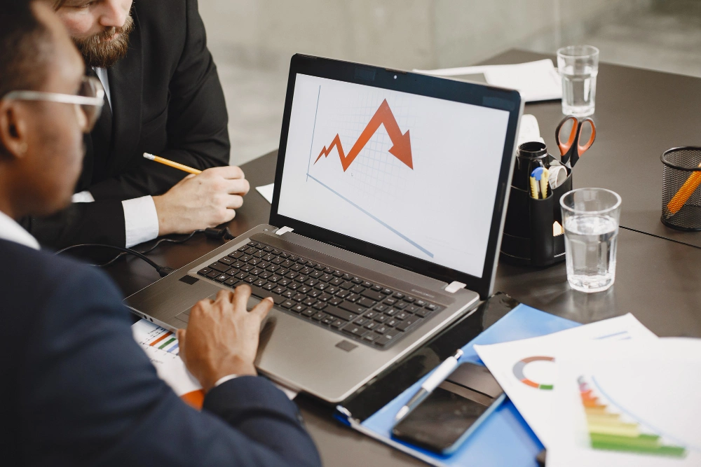Whissel Strategies Two people in business attire review a laptop displaying a line graph with red and orange sections, seated at a desk with documents, glasses of water, and office supplies. Toronto Digital Marketing Agency
