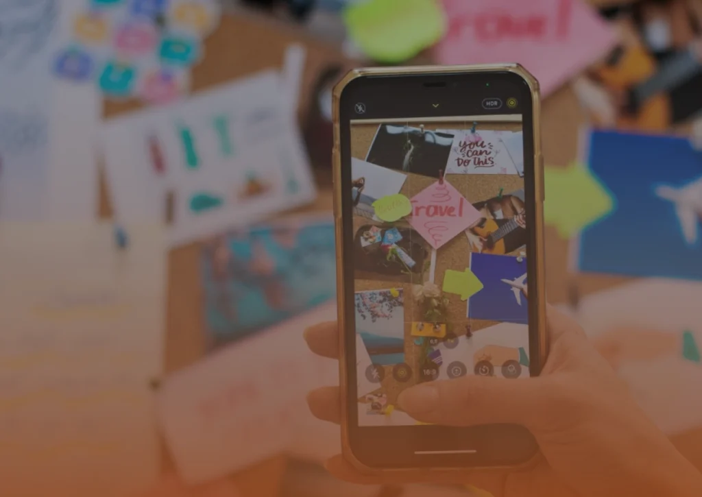 Whissel Strategies A hand holds a smartphone, taking a photo of a vision board with travel images, sticky notes, and motivational quotes on a cluttered table. Toronto Digital Marketing Agency